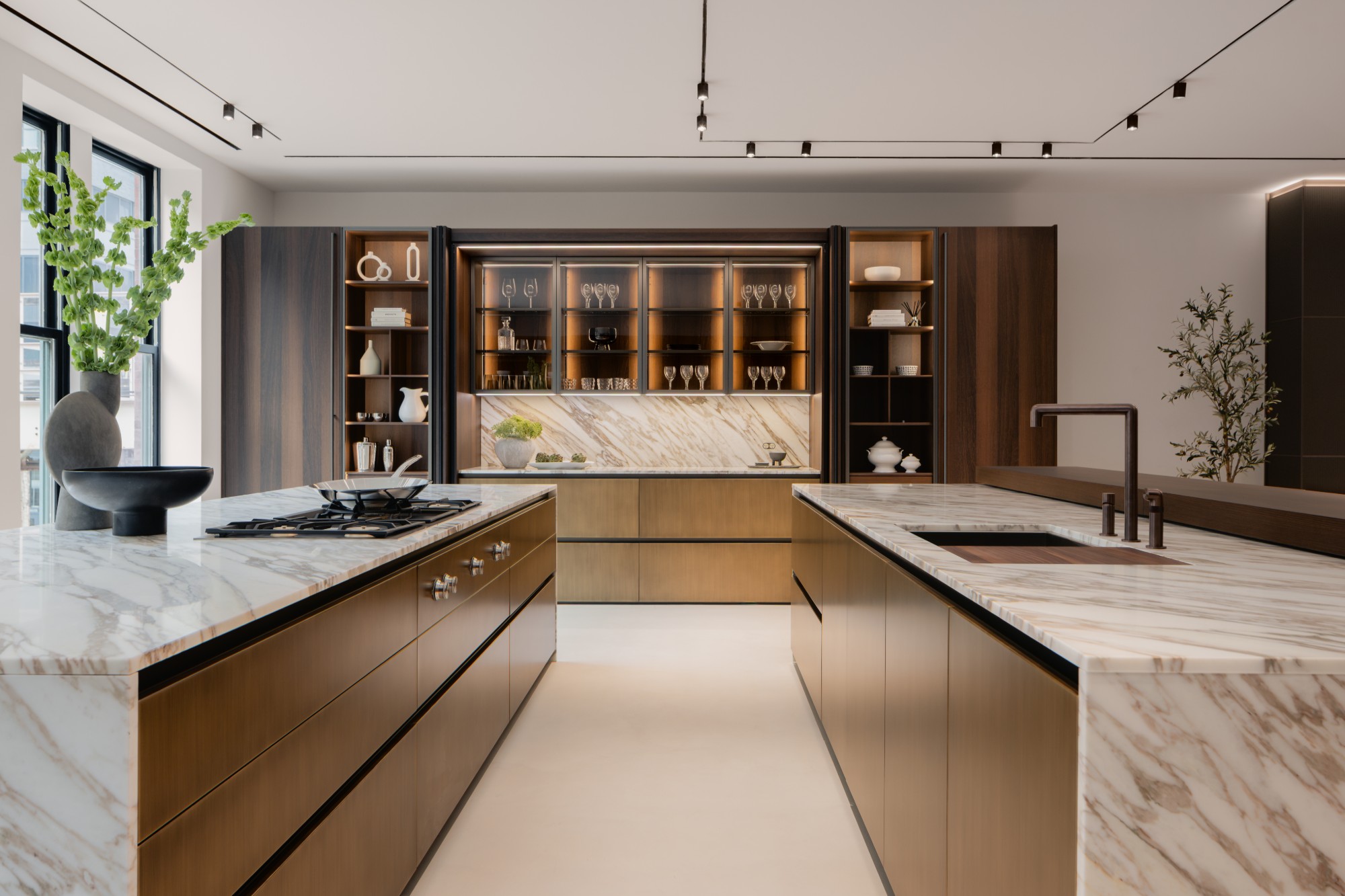 Two kitchen islands in marble and bronze before a lit glass cabinet wall